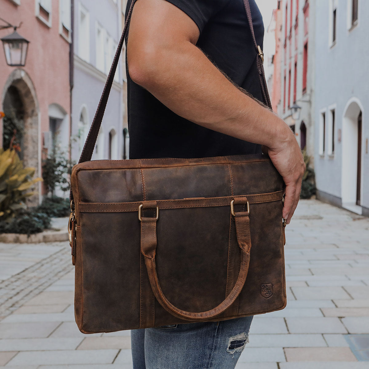 Person trägt eine braune Leder-Laptoptasche mit Schulterriemen und Griffen über der Schulter, steht auf einer gepflasterten Straße mit bunten Häusern im Hintergrund