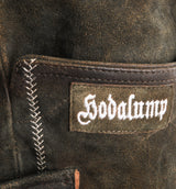 Detailaufnahme einer traditionellen Hodalump-Lederhose aus dunklem Wildleder mit weißer Ziernaht und aufgesticktem Hodalump-Logo.