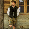 Person lehnt an einer Holzwand, trägt traditionelle bayerische Kleidung mit weißem Hemd, grüner Weste und brauner Lederhose