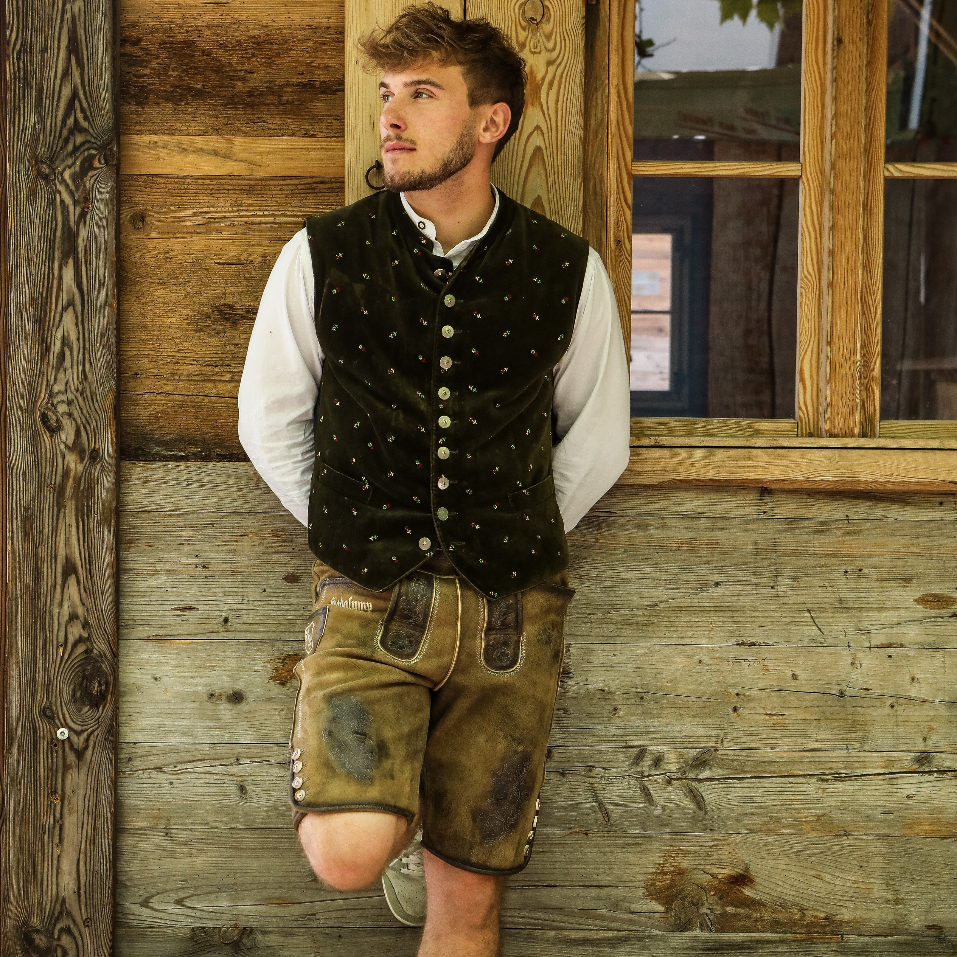 Person lehnt an einer Holzwand, trägt traditionelle bayerische Kleidung mit weißem Hemd, grüner Weste und brauner Lederhose