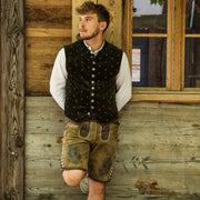 Person lehnt an einer Holzwand, trägt traditionelle bayerische Kleidung mit weißem Hemd, grüner Weste und brauner Lederhose