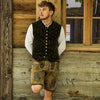 Person lehnt an einer Holzwand, trägt traditionelle bayerische Kleidung mit weißem Hemd, grüner Weste und brauner Lederhose