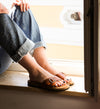 Person sitzt im Sonnenlicht auf einer Fensterbank, trägt hochgekrempelte Jeans und braune Ledersandalen mit silberner Schnalle.