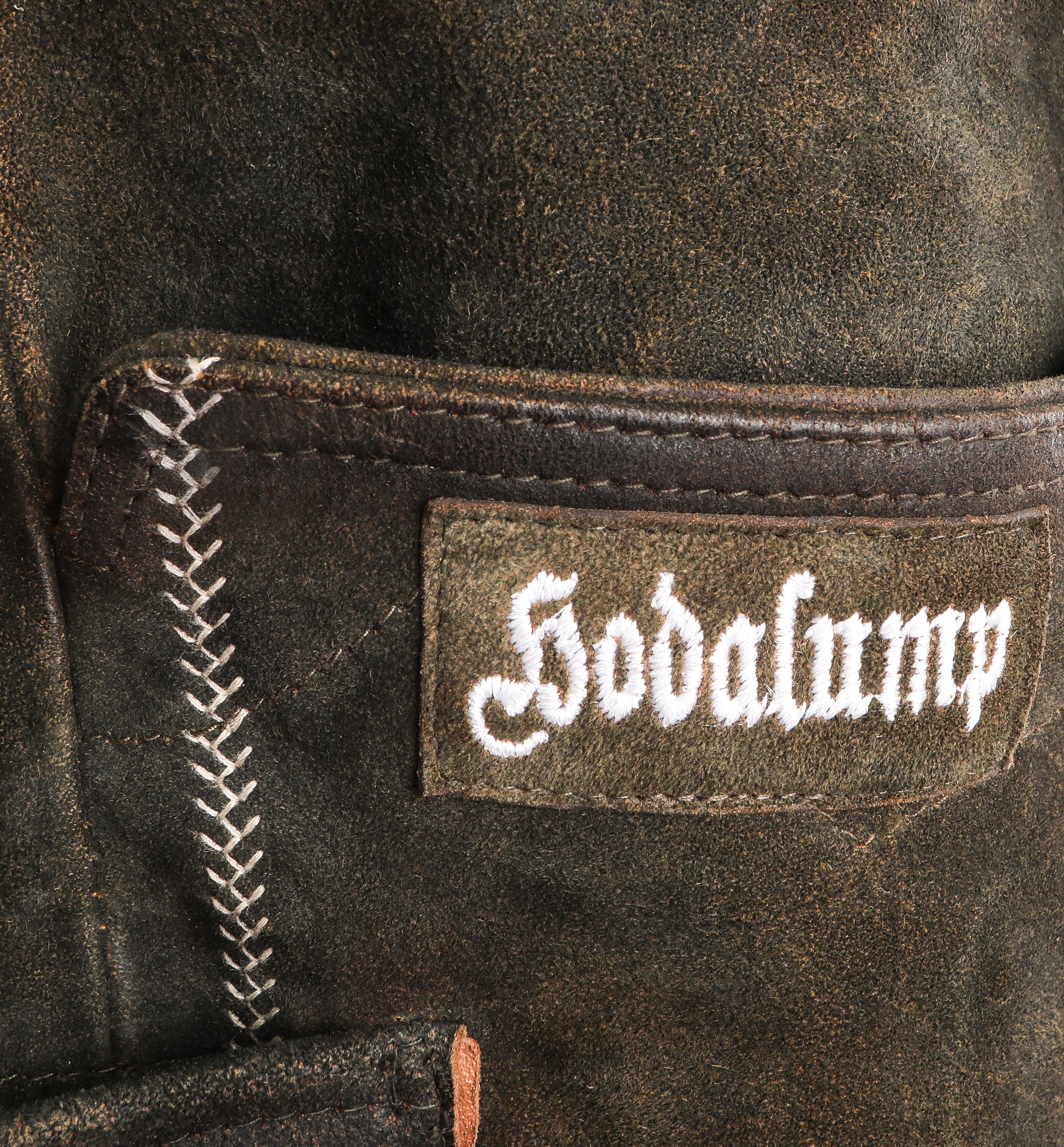 Detailaufnahme einer traditionellen Hodalump-Lederhose aus dunklem Wildleder mit weißer Ziernaht und aufgesticktem Hodalump-Logo.