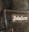 Detailaufnahme einer traditionellen Hodalump-Lederhose aus dunklem Wildleder mit weißer Ziernaht und aufgesticktem Hodalump-Logo.