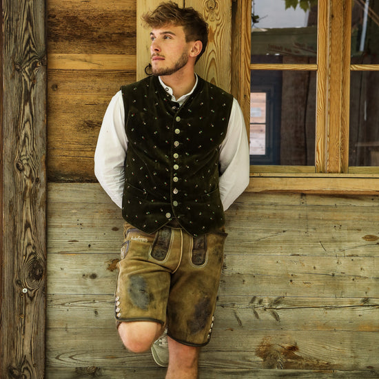 Person lehnt an einer Holzwand, trägt traditionelle bayerische Kleidung mit weißem Hemd, grüner Weste und brauner Lederhose