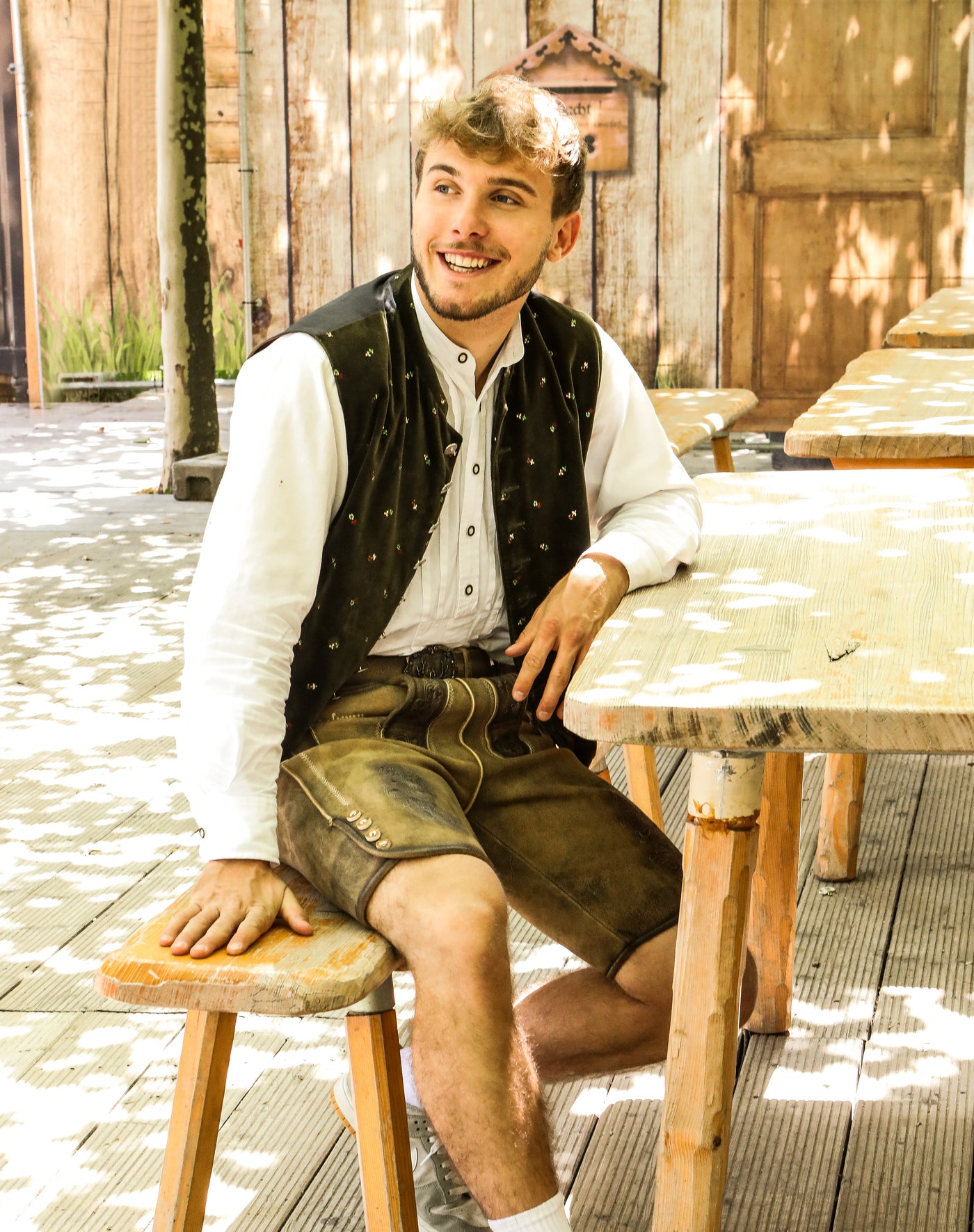 Der Mann sitzt entspannt auf einer Holzbank und trägt eine traditionelle kurze Lederhose, ein weißes Trachtenhemd und eine bestickte schwarze Weste.