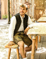 Der Mann sitzt entspannt auf einer Holzbank und trägt eine traditionelle kurze Lederhose, ein weißes Trachtenhemd und eine bestickte schwarze Weste.